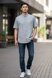 Man wearing a gray button-up shirt, blue jeans, and white sneakers standing on a sidewalk.