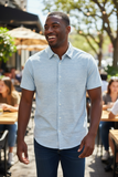 Man wearing a light blue button-up shirt standing outdoors with people and tables in the background