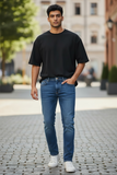 Man wearing a black t-shirt and blue jeans standing on a paved street.