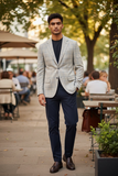 Man in a light gray blazer and dark blue pants standing outdoors in a casual setting.