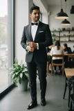 Man in a black tuxedo standing in a modern interior setting with people in the background.