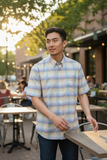 Man wearing a plaid shirt standing outdoors with tables and people in the background