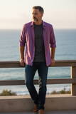 Man wearing a purple shirt and dark jeans standing by a wooden railing with ocean in the background