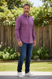 Man wearing a purple shirt and blue jeans standing outdoors with greenery in the background