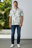 Man wearing a patterned shirt and jeans standing outdoors with plants in the background