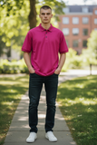 Person wearing a red polo shirt and dark jeans standing on a path with greenery and a building in the background.