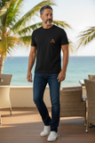 Man wearing a black t-shirt with a logo and blue jeans standing on a wooden deck with palm trees and ocean view.