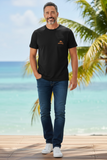 Man wearing a black t-shirt with a logo and blue jeans standing on a wooden deck with palm trees and ocean in the background.