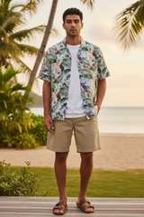 Man wearing a tropical floral shirt and beige shorts standing on a wooden deck with palm trees and ocean in the background.