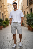 Man wearing a white t-shirt and light gray shorts standing on a stone street with plants and buildings in the background.