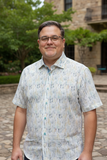 Man wearing glasses and a patterned shirt standing outdoors with greenery and a building in the background