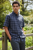 Man wearing a blue plaid shirt standing outdoors with greenery in the background