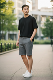 Man wearing a black t-shirt and gray shorts standing on a sidewalk with trees and buildings in the background.