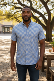 Man wearing a blue patterned shirt standing outdoors with trees and a table in the background