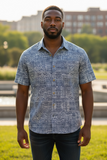 Man wearing a blue patterned shirt standing outdoors with greenery and buildings in the background