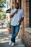 Man standing against a brick wall wearing a light blue shirt and jeans.